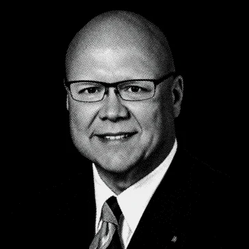 Man in a suit and tie wearing glasses, smiling slightly, posed for a professional portrait against a plain background.
